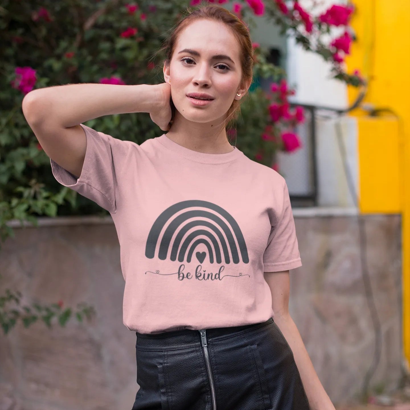Woman wearing a pink t-shirt with a rainbow design and 'be kind' text, standing outdoors with flowers in the background.