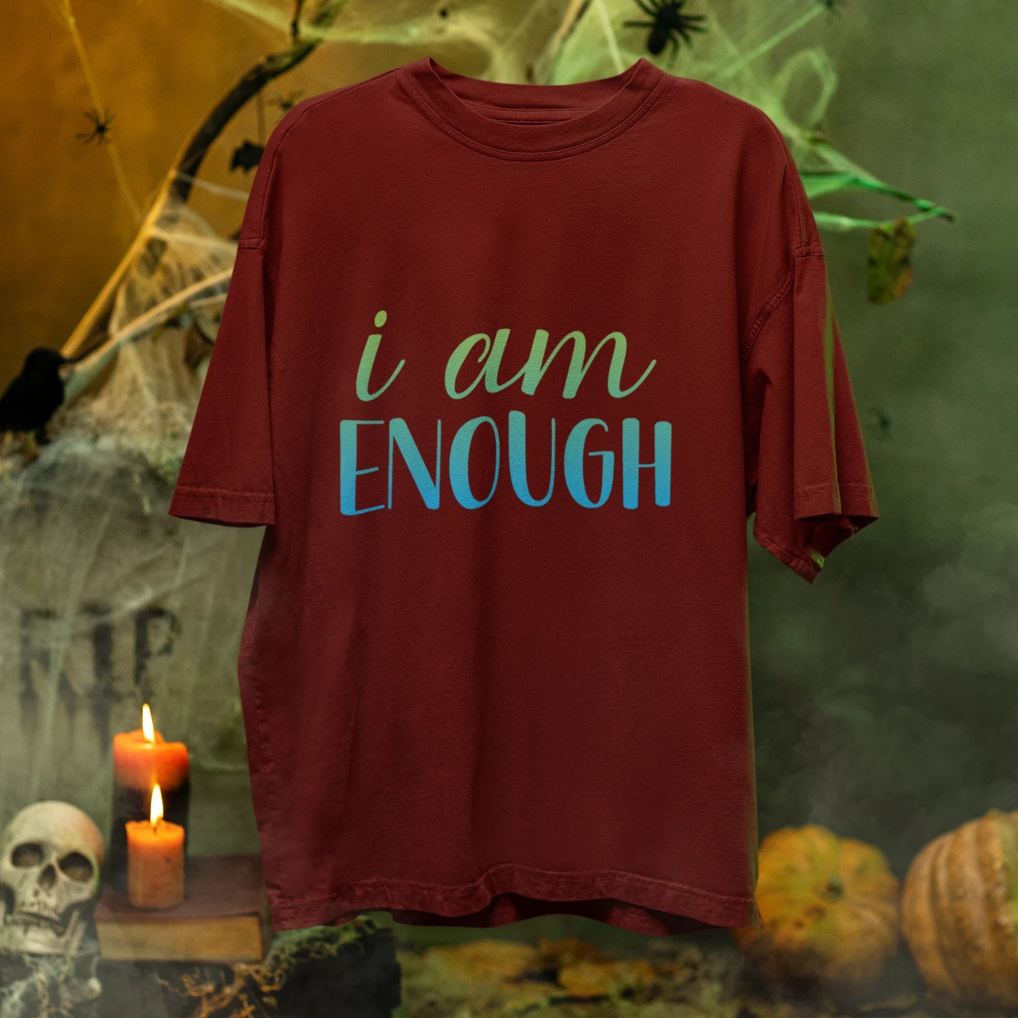 Red t-shirt with 'i am ENOUGH' text in a Halloween-themed setting with pumpkins, candles, and cobwebs.