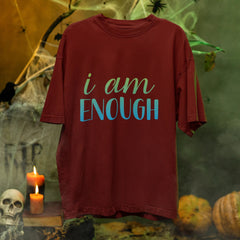 Red t-shirt with 'i am ENOUGH' text in a Halloween-themed setting with pumpkins, candles, and cobwebs.