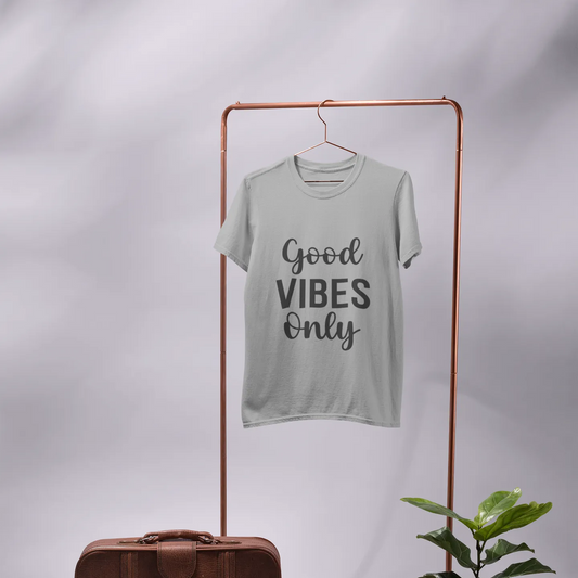 Light gray t-shirt with 'Good Vibes Only' text on a clothing rack with a suitcase, mirror, and plant.