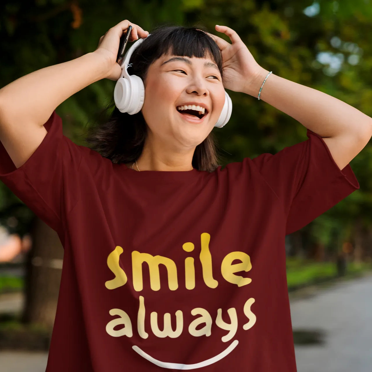 Person wearing a maroon t-shirt with 'smile always' text, holding white headphones, outdoors.