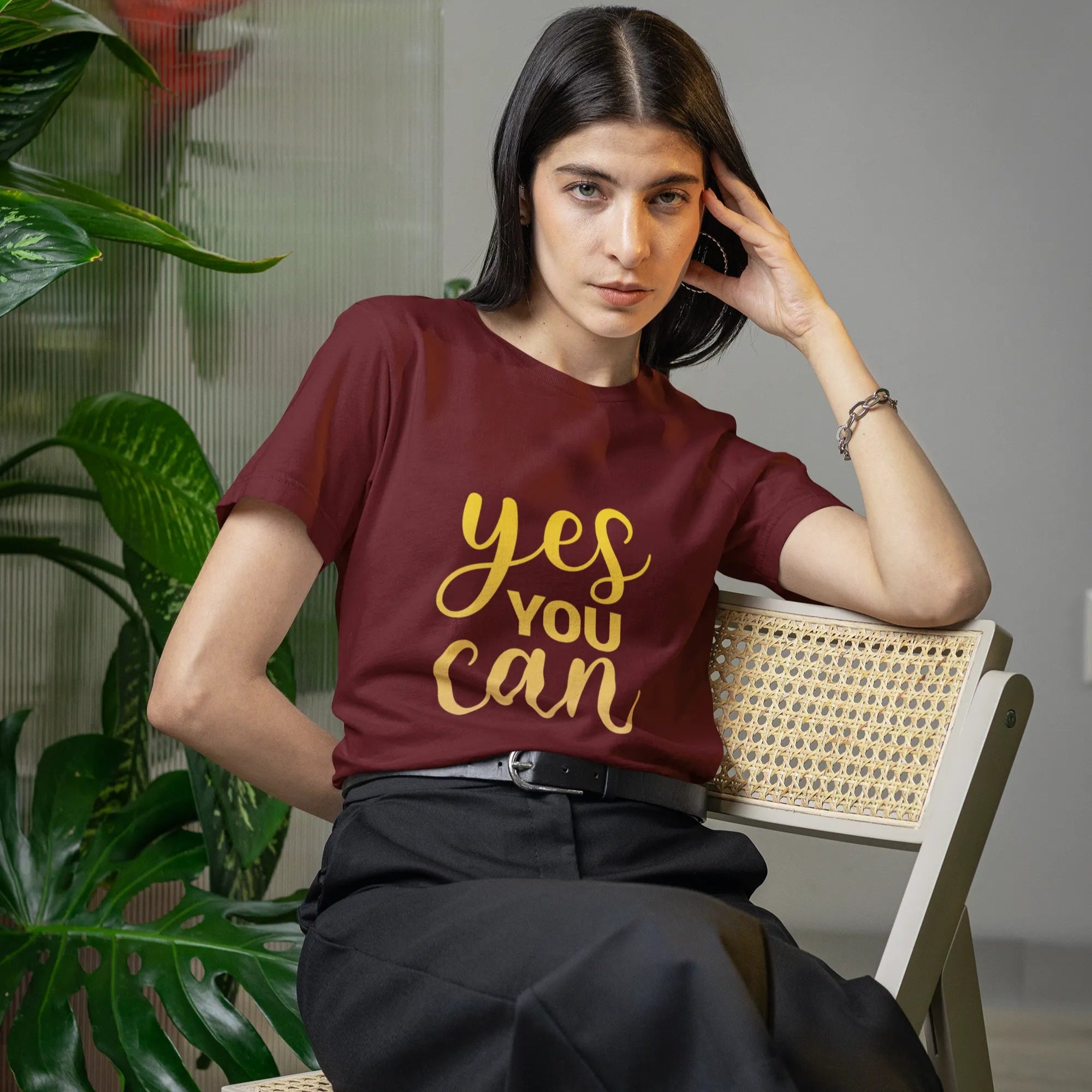 Woman wearing a maroon t-shirt with 'yes you can' in gold, sitting on a chair indoors.