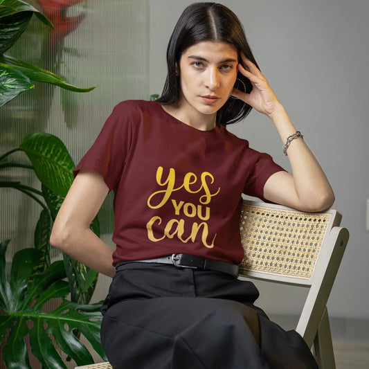 Woman wearing a maroon t-shirt with 'yes you can' in gold, sitting on a chair indoors.