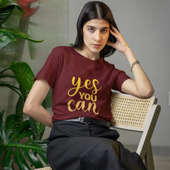Woman wearing a maroon t-shirt with 'yes you can' in gold, sitting on a chair indoors.