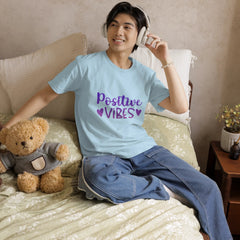 Person wearing a 'Positive Vibes' t-shirt sitting on a bed with a teddy bear.