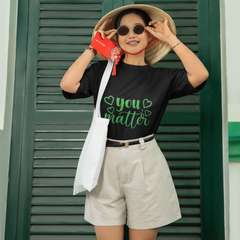 Person wearing a black t-shirt with 'you matter' text, beige shorts, and a wide-brimmed hat in front of green shutters with red lanterns.