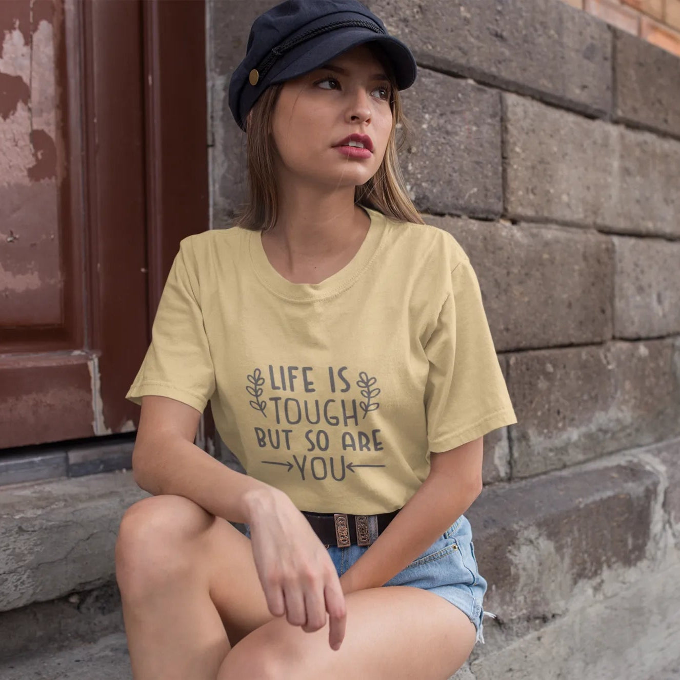 Woman wearing a yellow t-shirt with 'Life is tough but so are you' text, sitting against a brick wall.