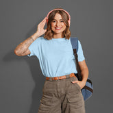 Woman wearing pink headphones, blue shirt, and brown pants against a gray background