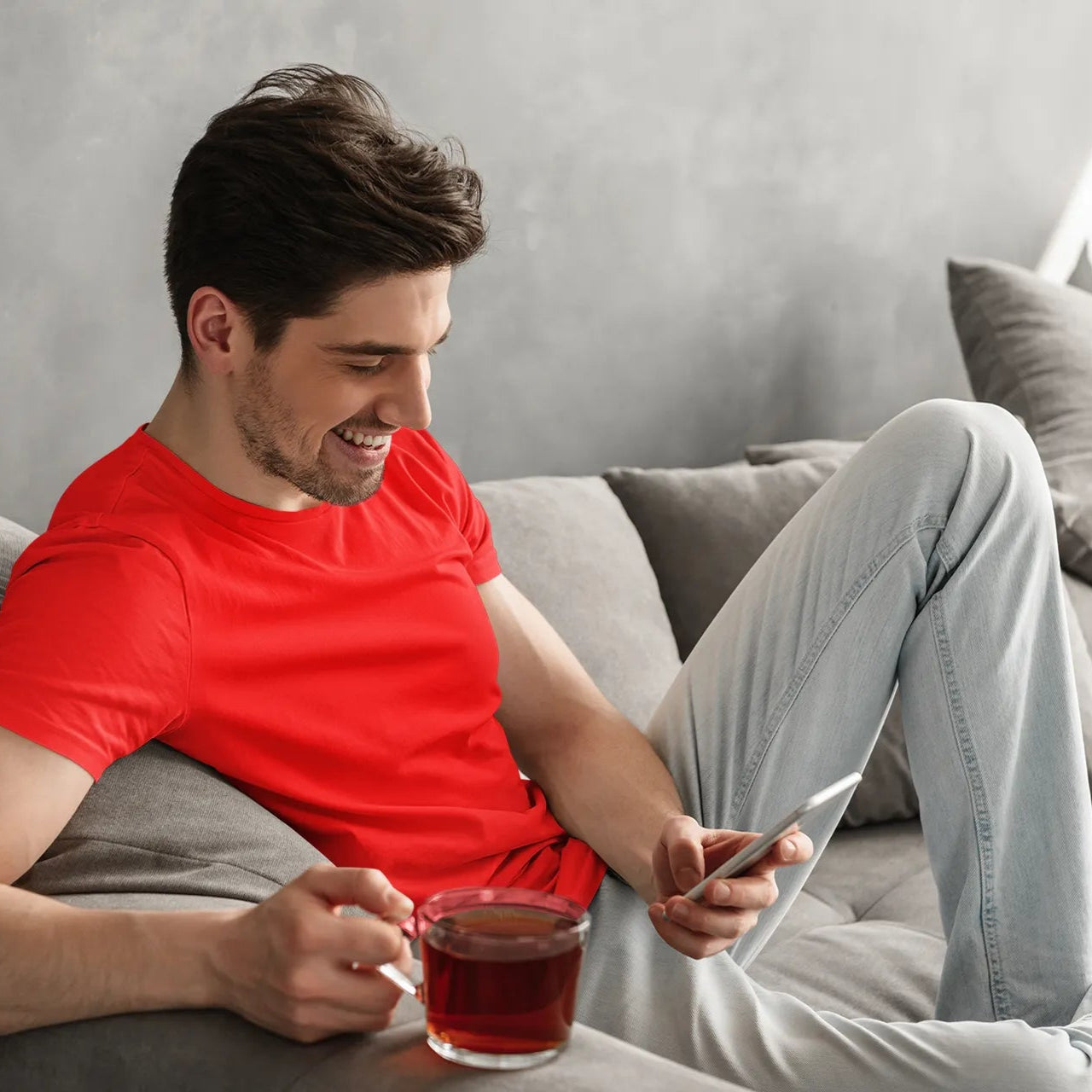 Man in red shirt sitting on a couch with a cup of tea, using a smartphone.