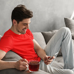 Man in red shirt sitting on a couch with a cup of tea, using a smartphone.