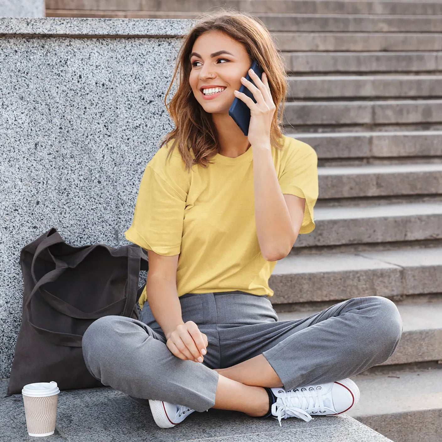 Woman sitting on steps talking on a phone with a coffee cup nearby