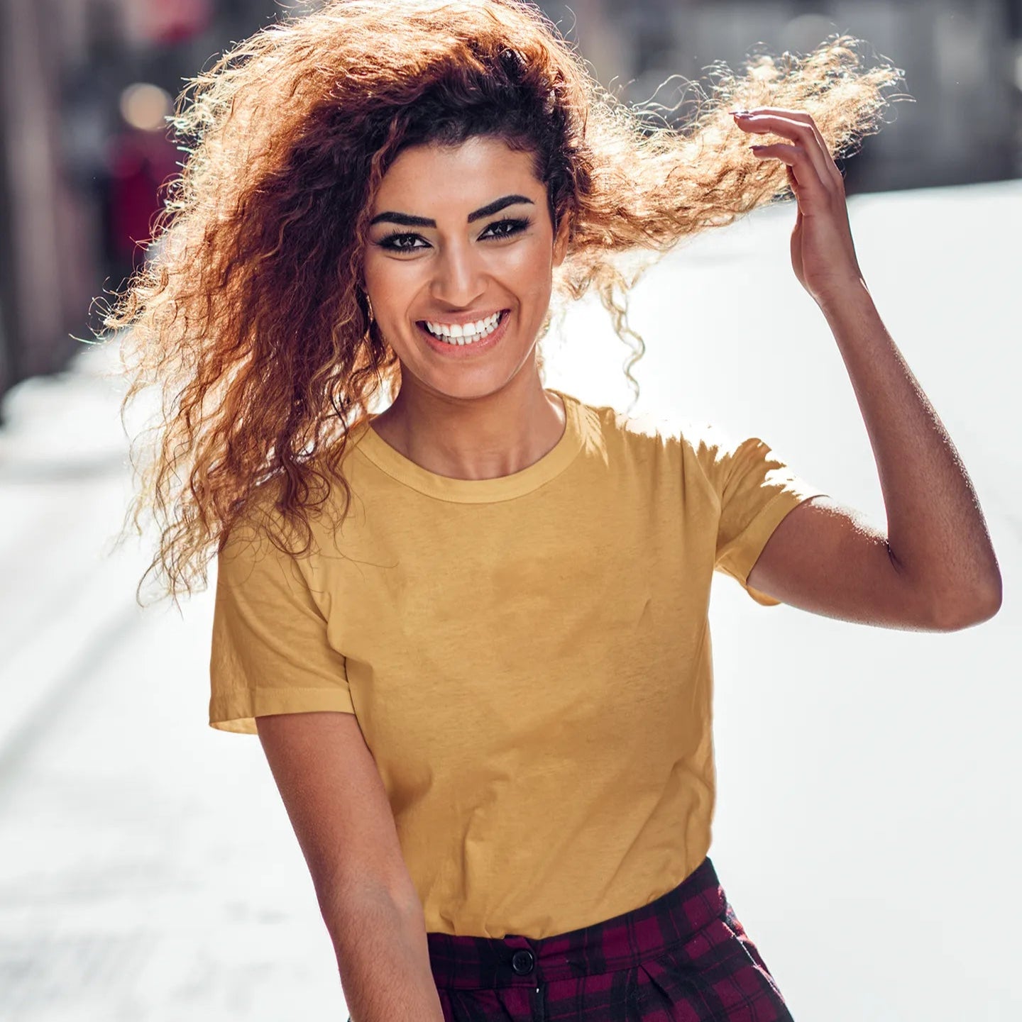 Woman with curly hair wearing a yellow shirt and plaid pants, smiling outdoors.