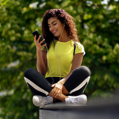 Woman sitting outdoors, using a phone with earphones, wearing a yellow top and black leggings.
