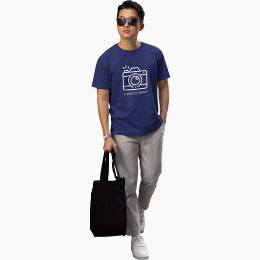 Person wearing a blue t-shirt with a camera graphic and text, holding a black tote bag on a white background