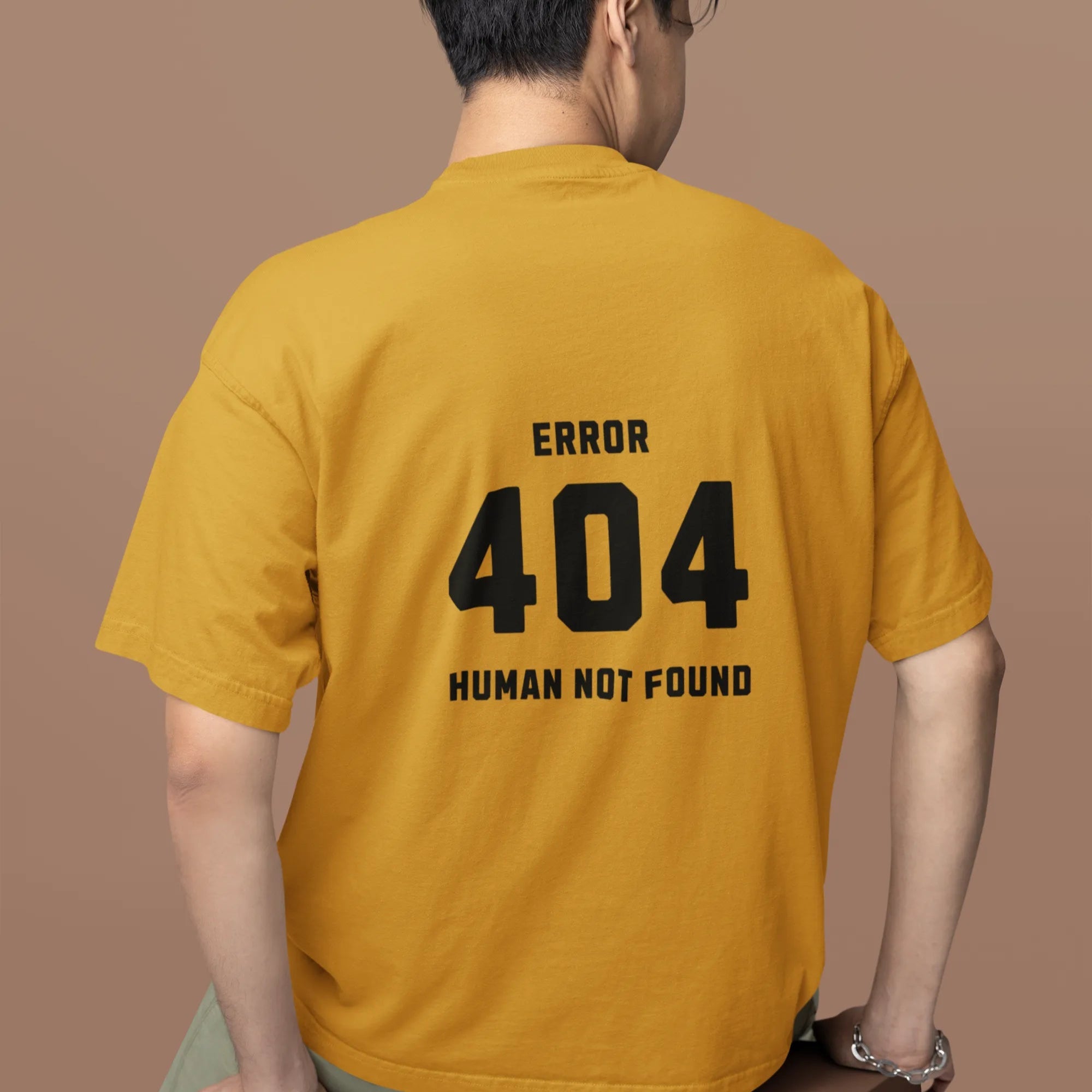 Person wearing a mustard yellow t-shirt with 'ERROR 404 HUMAN NOT FOUND' text on the back, sitting on a brown box against a brown background.