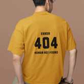 Person wearing a mustard yellow t-shirt with 'ERROR 404 HUMAN NOT FOUND' text on the back, sitting on a brown box against a brown background.