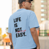 Person wearing a light blue t-shirt with 'Life is Not Easy' text on the back, standing outdoors.