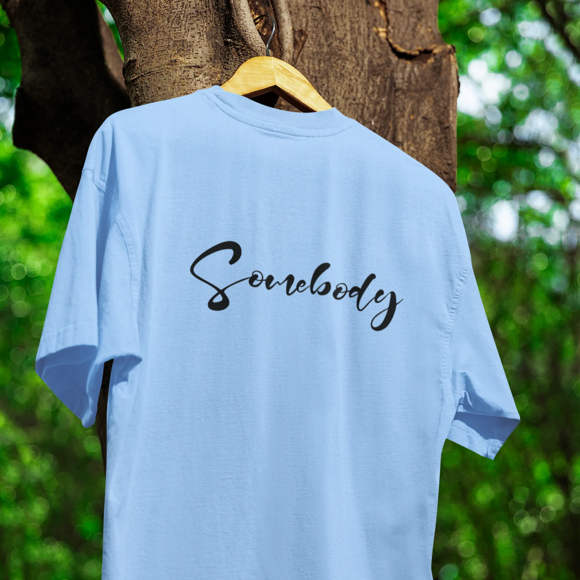 Light blue t-shirt with 'Somebody' text on a hanger against a natural background