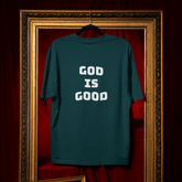 Teal t-shirt with 'GOD IS GOOD' text on a hanger against a red curtain background.
