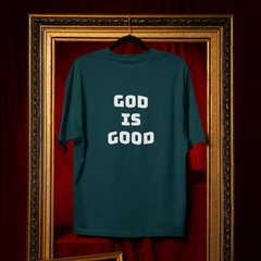 Teal t-shirt with 'GOD IS GOOD' text on a hanger against a red curtain background.