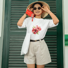 Woman wearing a white t-shirt with a floral design, beige shorts, and a wide-brimmed hat in front of green shutters with red lanterns.