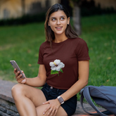 Woman sitting on a bench in a park, holding a phone and wearing a brown t-shirt with a flower design.