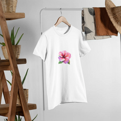White t-shirt with a pink flower design on a hanger against a white wall with a wooden shelf and plants.