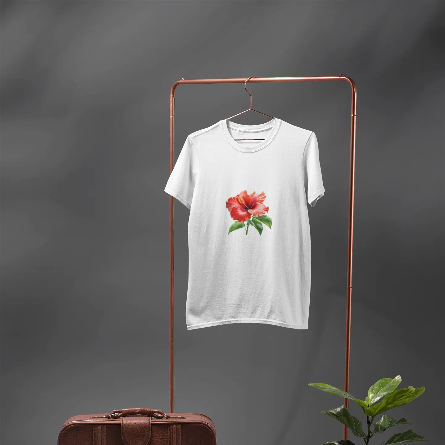 White t-shirt with a red flower design on a copper stand against a gray background, with a brown leather suitcase and potted plant.