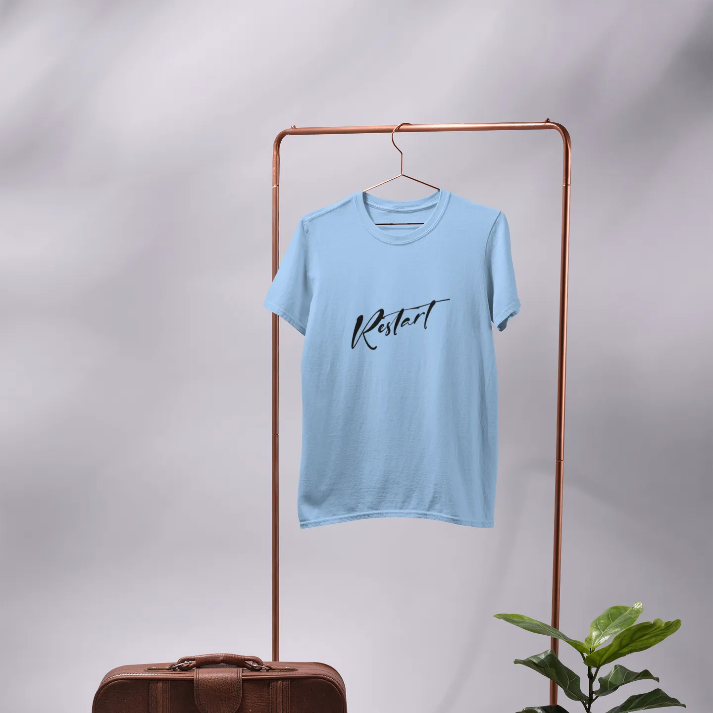 Light blue t-shirt with 'Kermit' text on a hanger, next to a brown suitcase and potted plant on a gray background