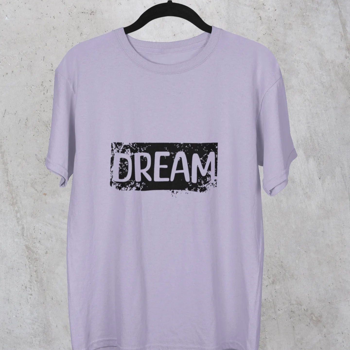 Lavender t-shirt with 'DREAM' print on a hanger against a gray concrete wall.
