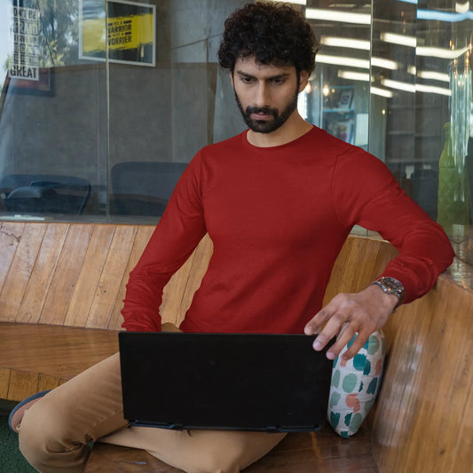 Man in a red shirt sitting on a wooden bench with a laptop, in an indoor setting.
