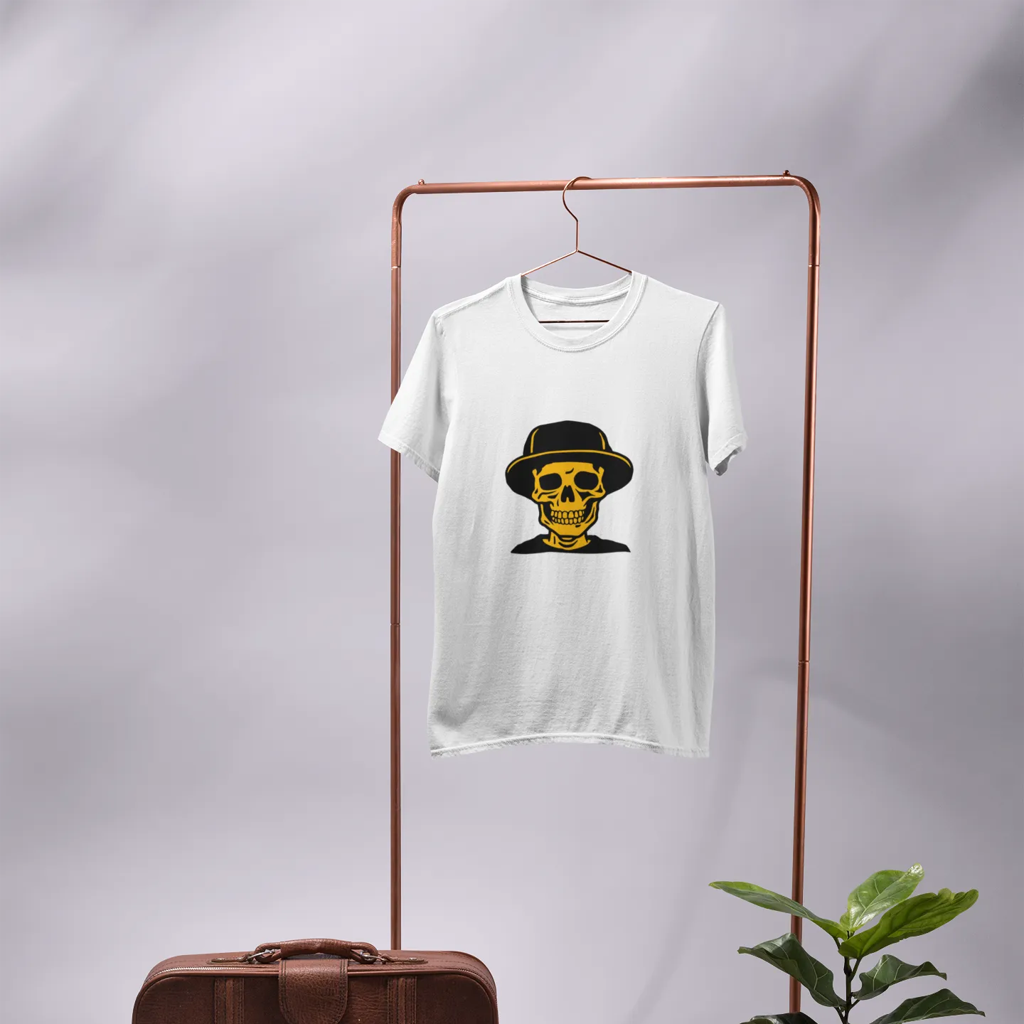 White t-shirt with a graphic design on a rack, next to a brown suitcase and potted plant.