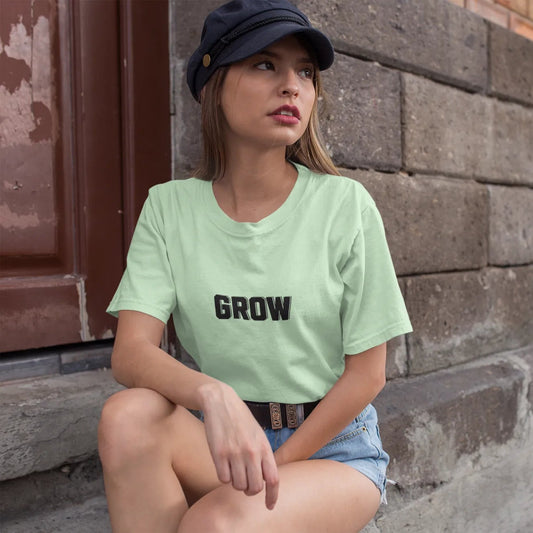 Woman wearing a light green t-shirt with 'GROW' printed on it, sitting against a brick wall.