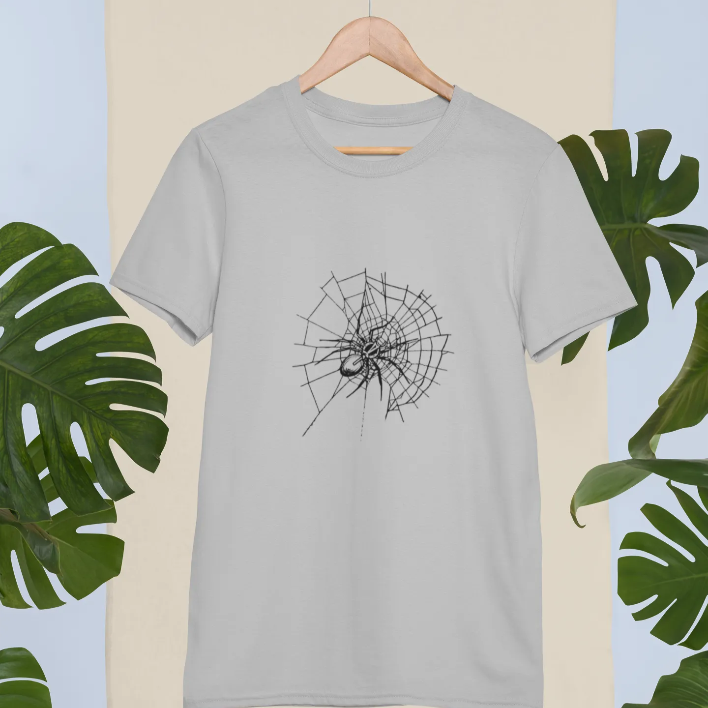 Gray t-shirt with black spider web design on a beige background with green leaves.