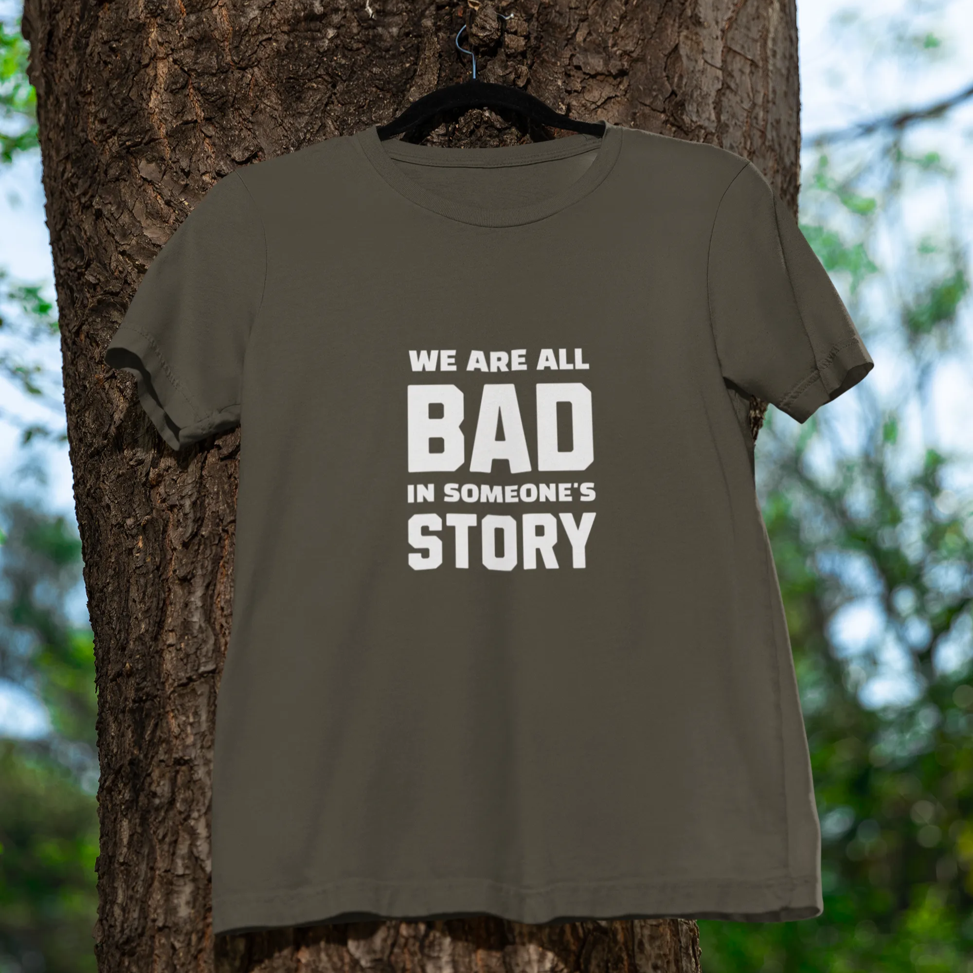 Green t-shirt with text 'WE ARE ALL BAD IN SOMEONE'S STORY' hanging on a tree