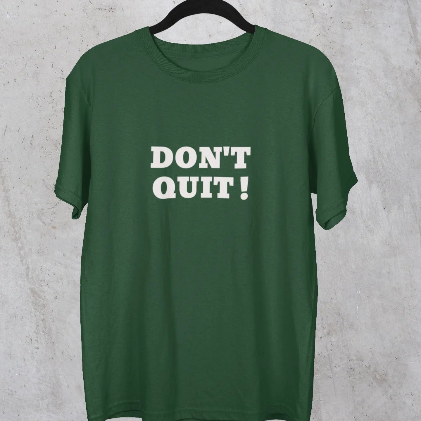 Green t-shirt with 'DON'T QUIT!' text on a hanger against a gray concrete wall.
