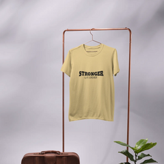 Yellow t-shirt with 'STRONGER' text on a hanger, next to a brown leather suitcase and potted plant against a gray background.