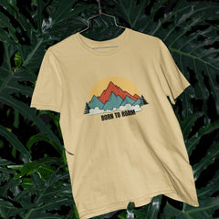 Beige t-shirt with mountain graphic and 'Born to Romm' text on a hanger against a leafy background