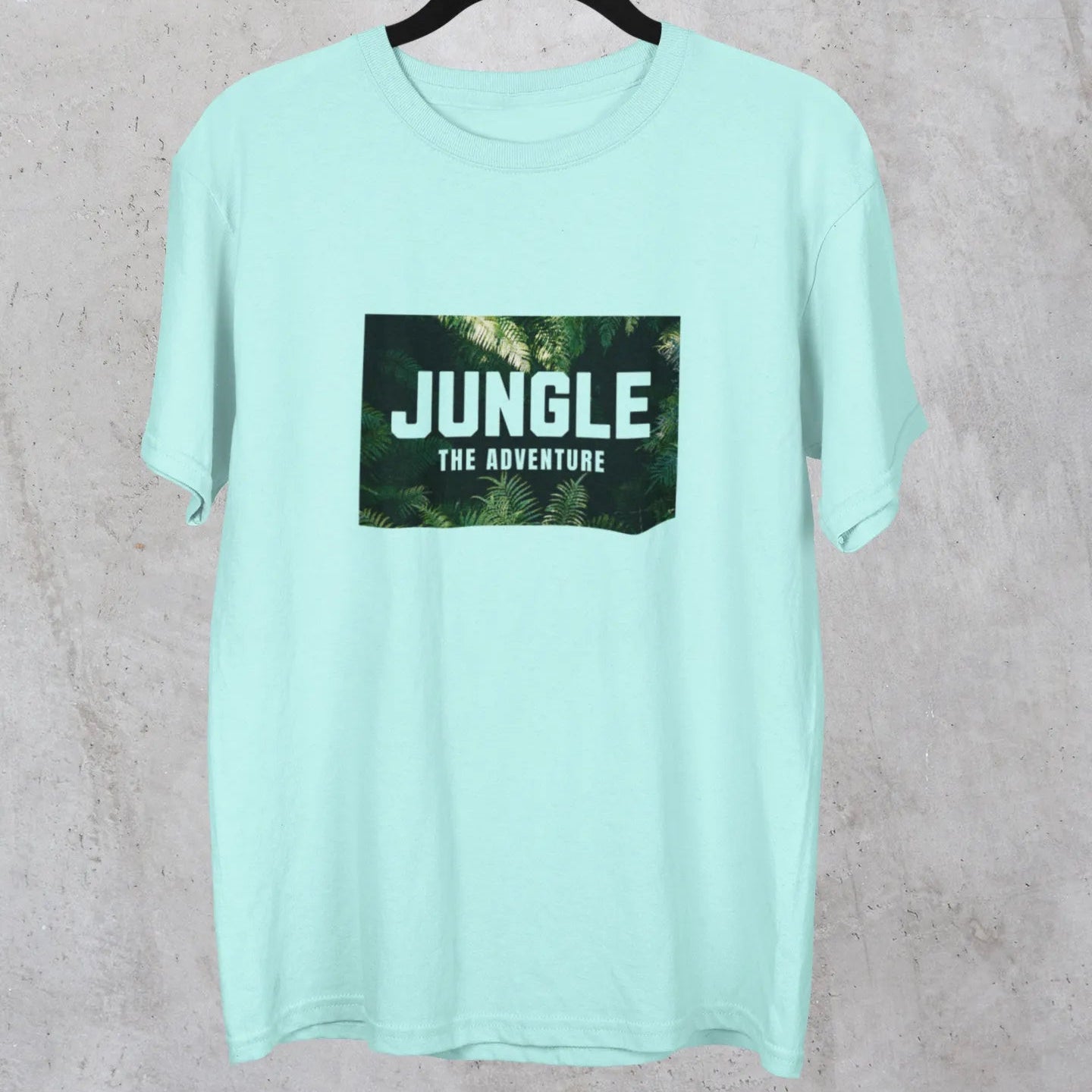 Mint green t-shirt with 'Jungle The Adventure' print on a hanger against a gray background