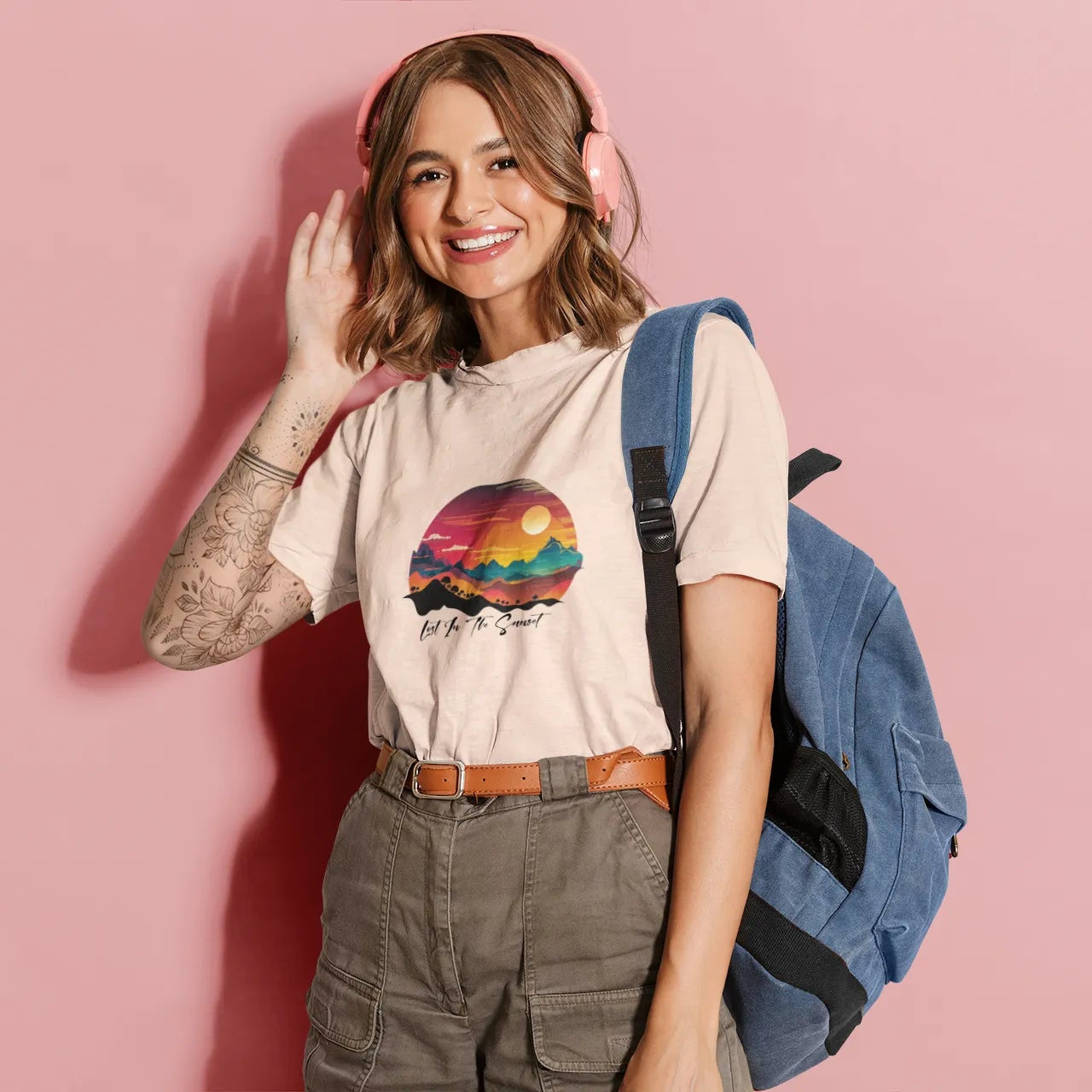 Woman wearing a t-shirt with a colorful design, holding headphones, and standing against a pink background.