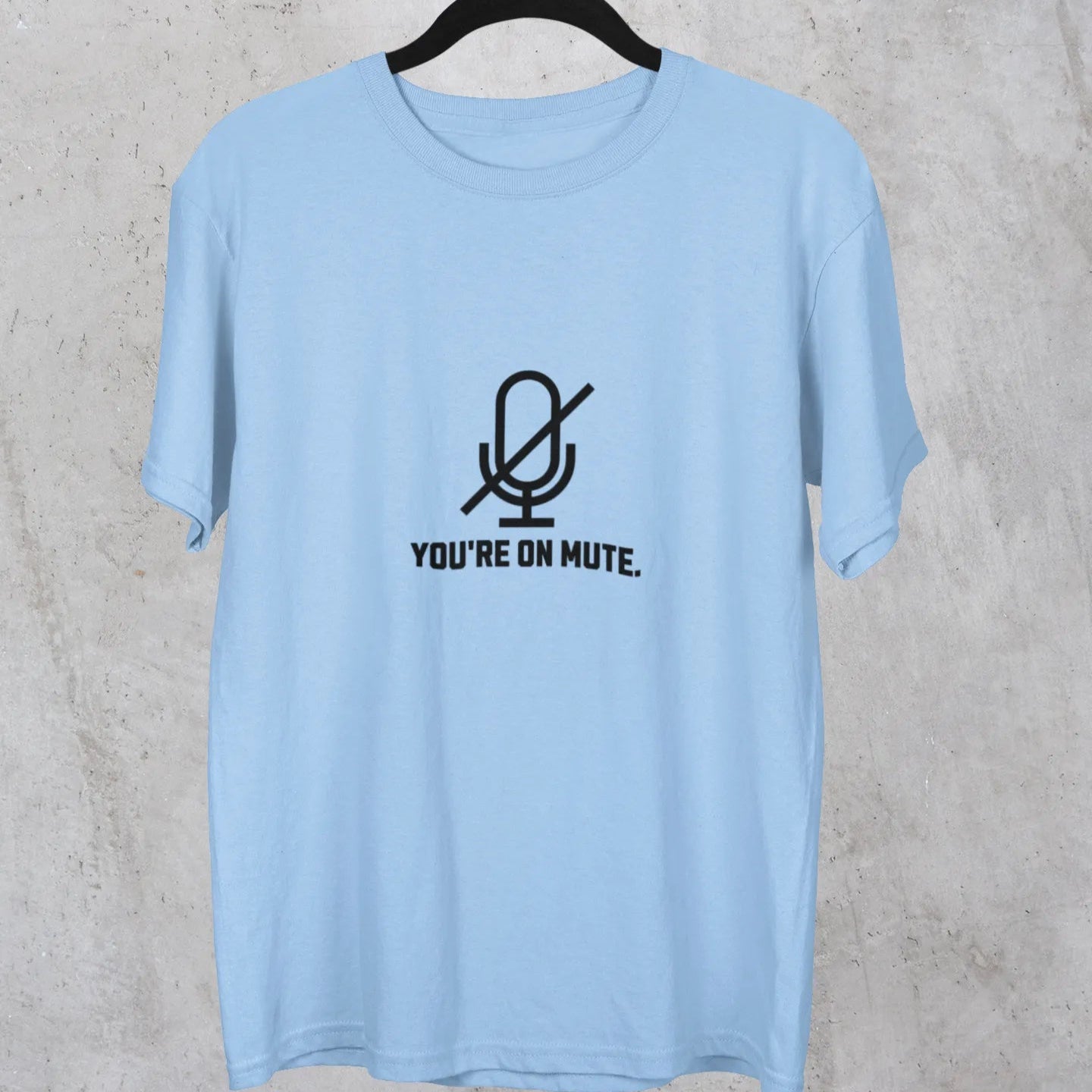 Light blue t-shirt with a microphone graphic and text on a hanger against a concrete wall.
