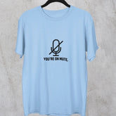 Light blue t-shirt with a microphone graphic and text on a hanger against a concrete wall.
