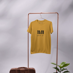Yellow t-shirt with black text on a hanger, next to a brown suitcase and potted plant on a gray background