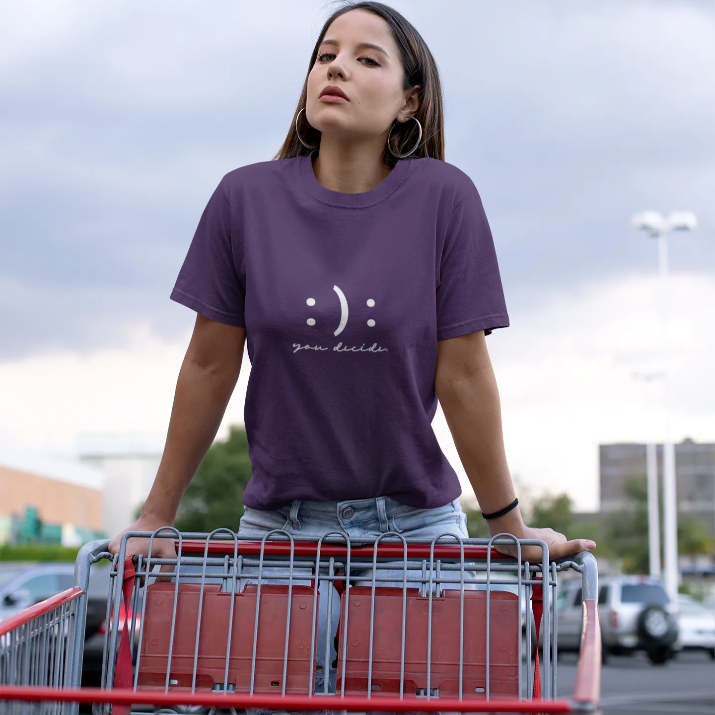 Woman wearing a purple t-shirt with a smiley face design, standing next to a shopping cart in an outdoor setting.