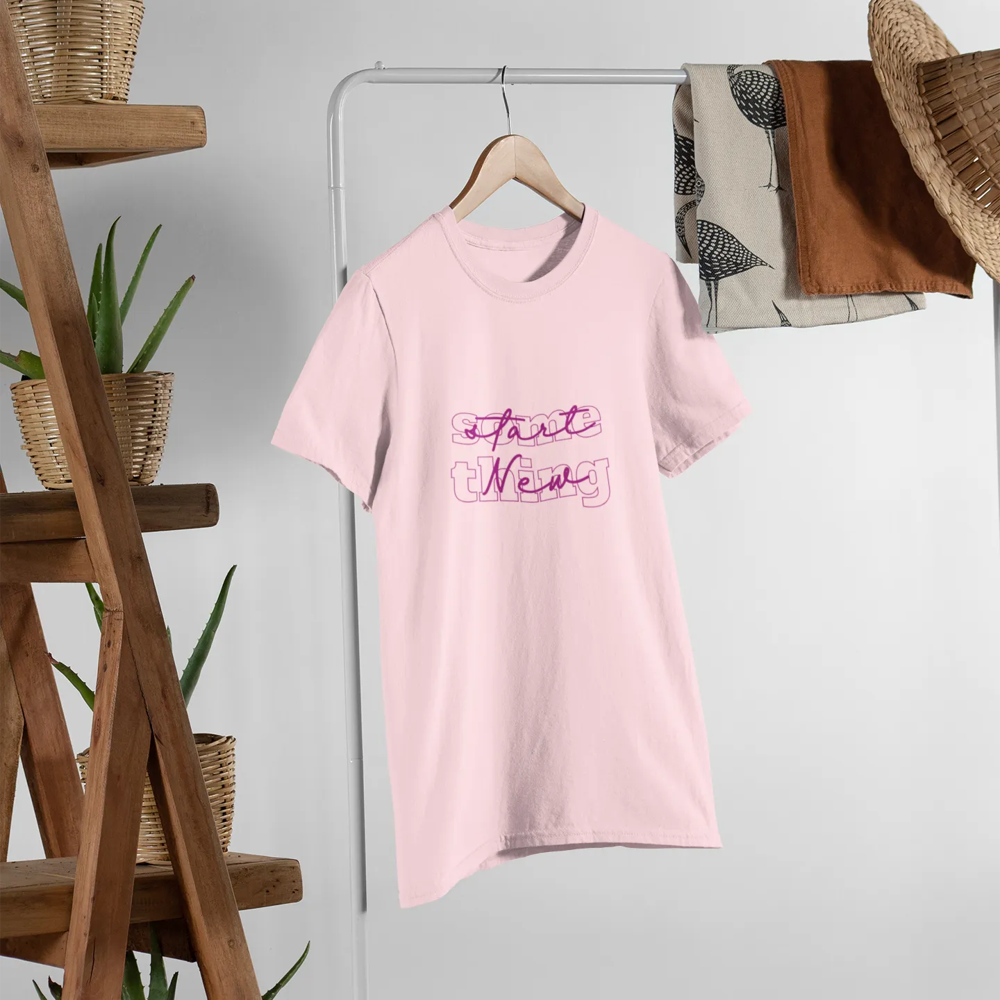Pink t-shirt with text on a hanger against a white wall with wooden shelves and plants.