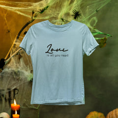 White t-shirt with 'Love is all you need' text on a Halloween-themed background with pumpkins, candles, and cobwebs.