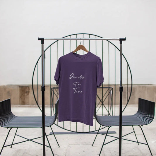Purple t-shirt with text on a black metal stand against a white wall.