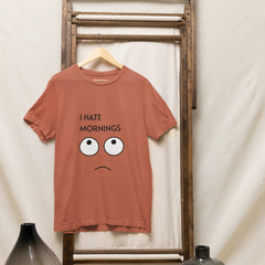 Brown t-shirt with 'I Hate Mornings' text and cartoon face on a wooden rack against a white curtain.