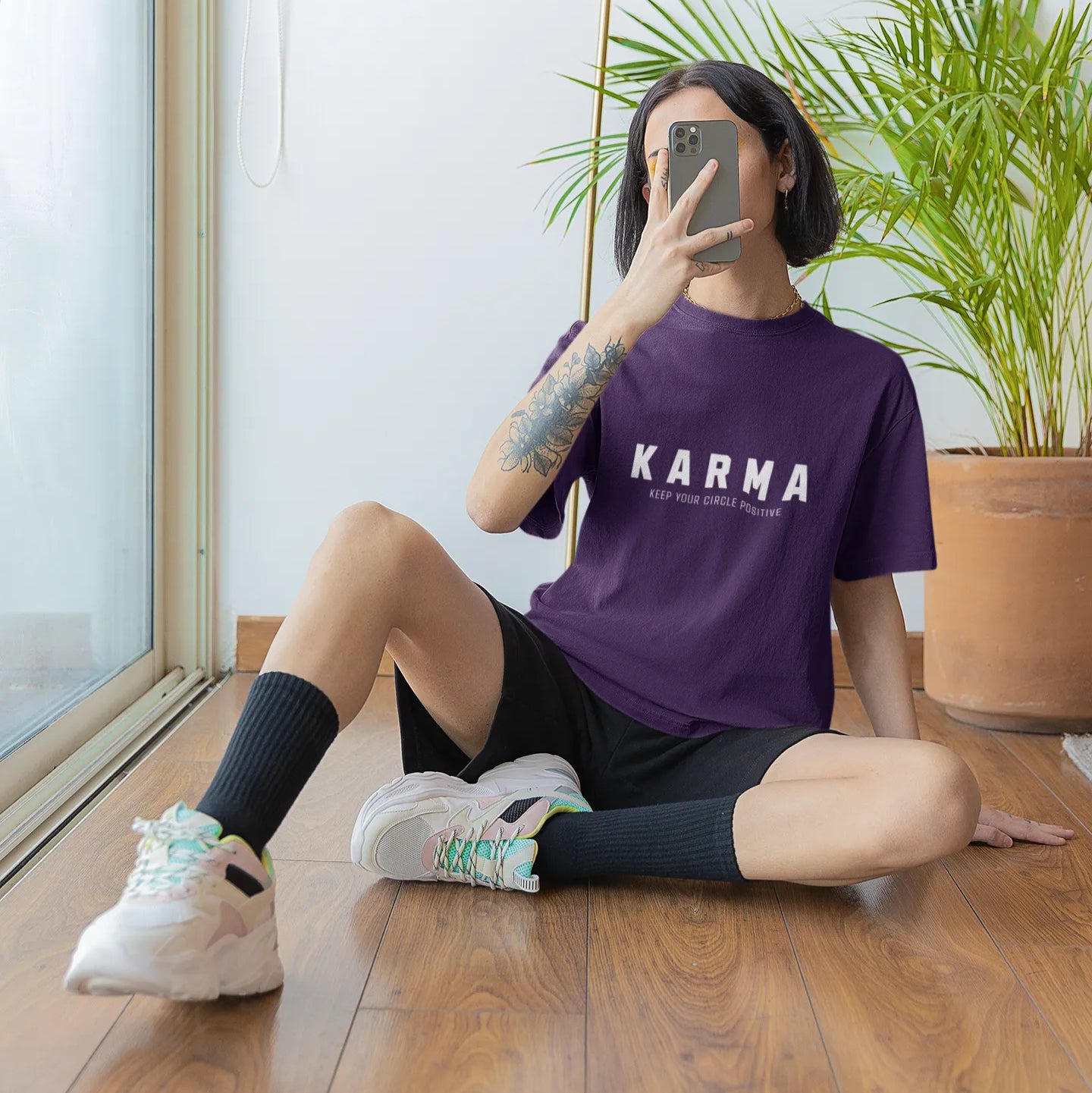 Person wearing a purple 'Karma' t-shirt sitting on the floor with plants and a lamp in the background.