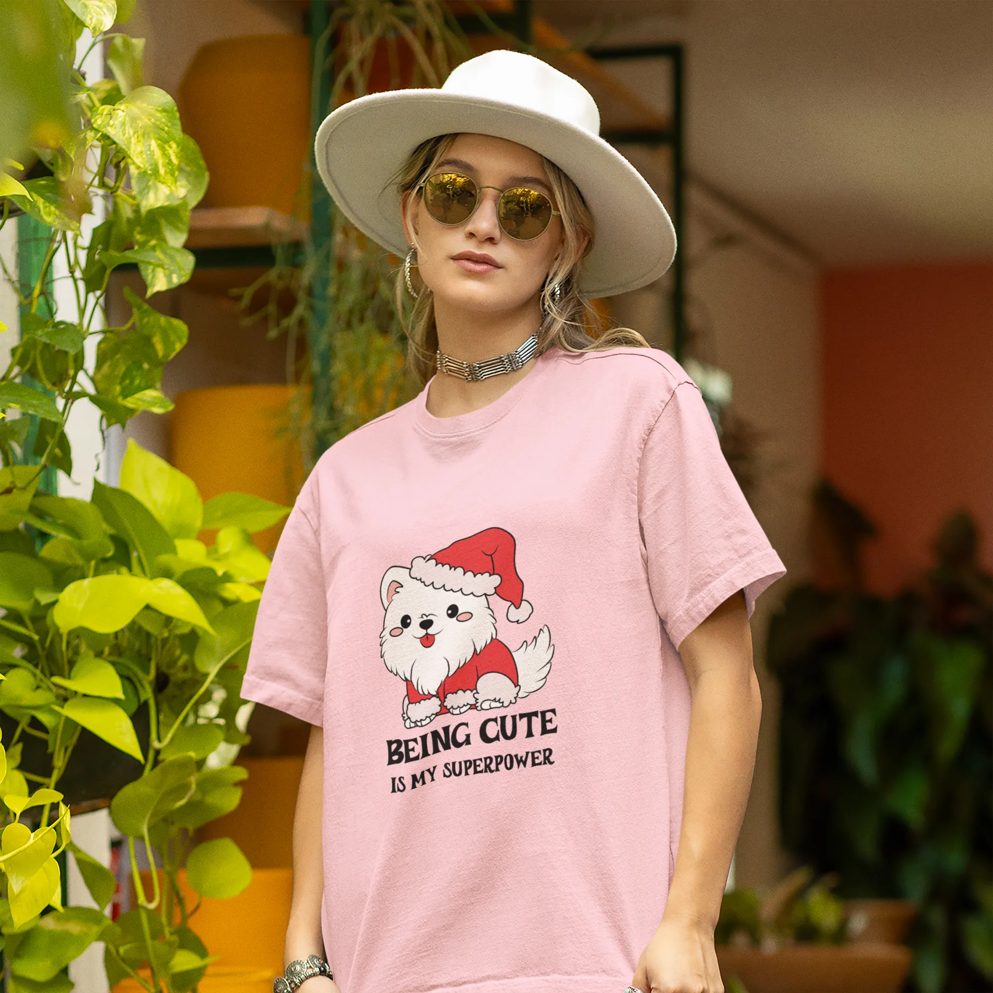 Person wearing a pink t-shirt with a cartoon dog in a Santa hat and text, standing in an outdoor setting with plants.